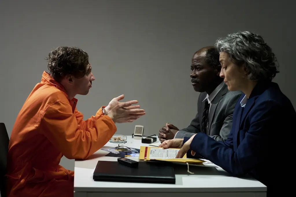Man wearing an orange prison jumpsuit sitting across a table during an interview.